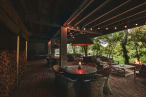 the garrison patio sits in a shaded area lit by lantern and lights. wicker chairs and thick wood tables