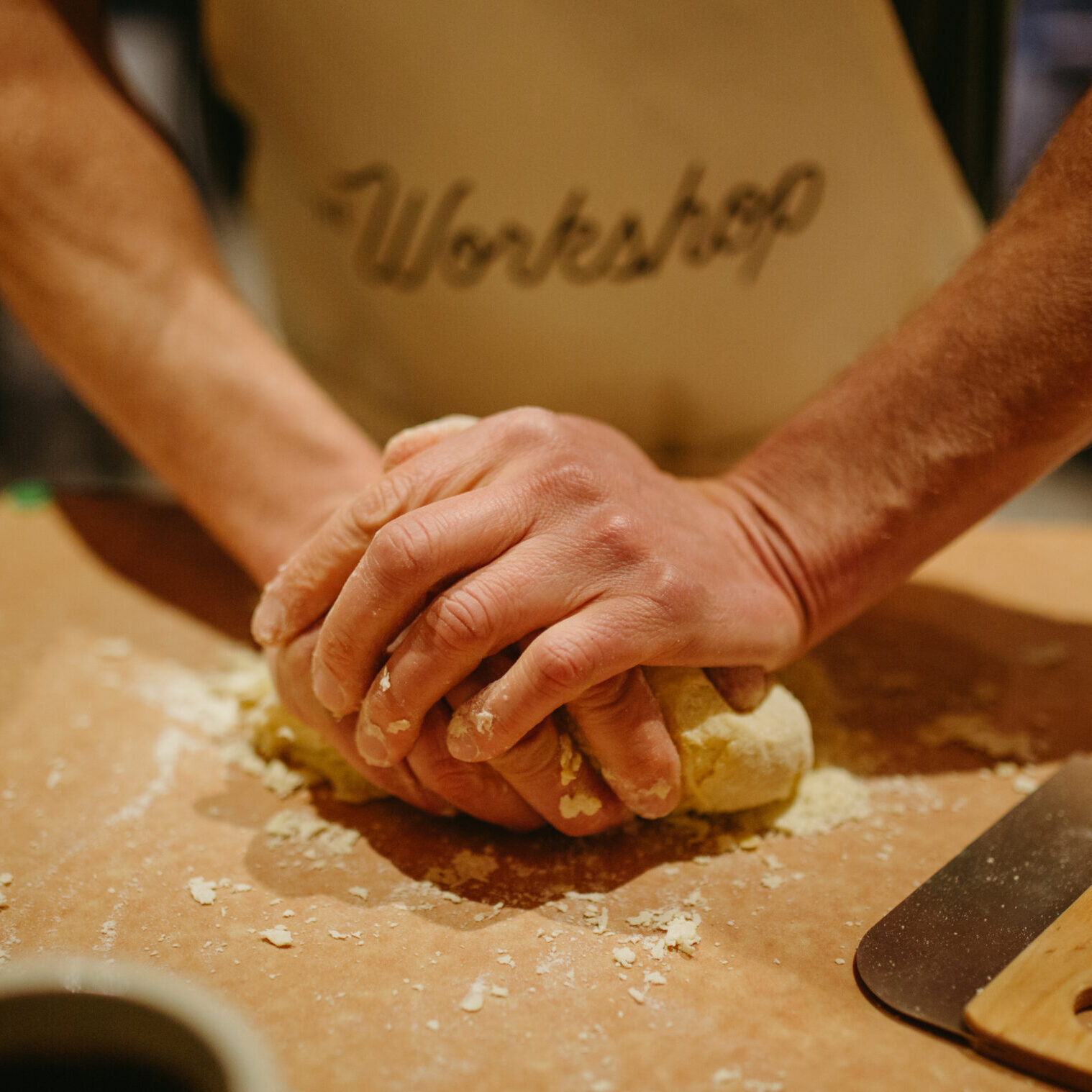 Handmade-Pasta-Class-The-Workshop (1) a man kneads a large piece of dough on a wooden board with flour sprinkled upon it
